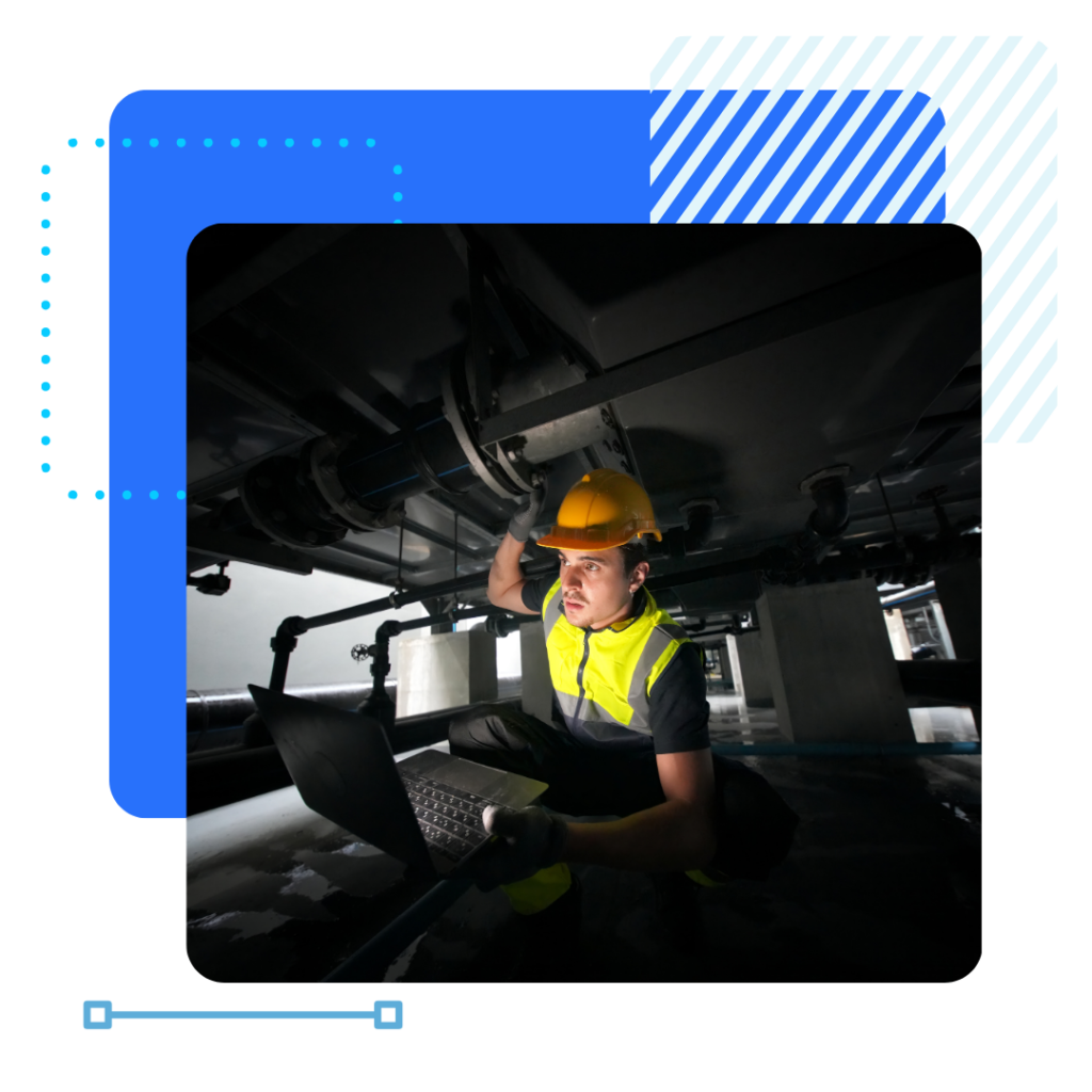 A facility management worker in a yellow vest and hard hat inspects machinery with a laptop in hand.