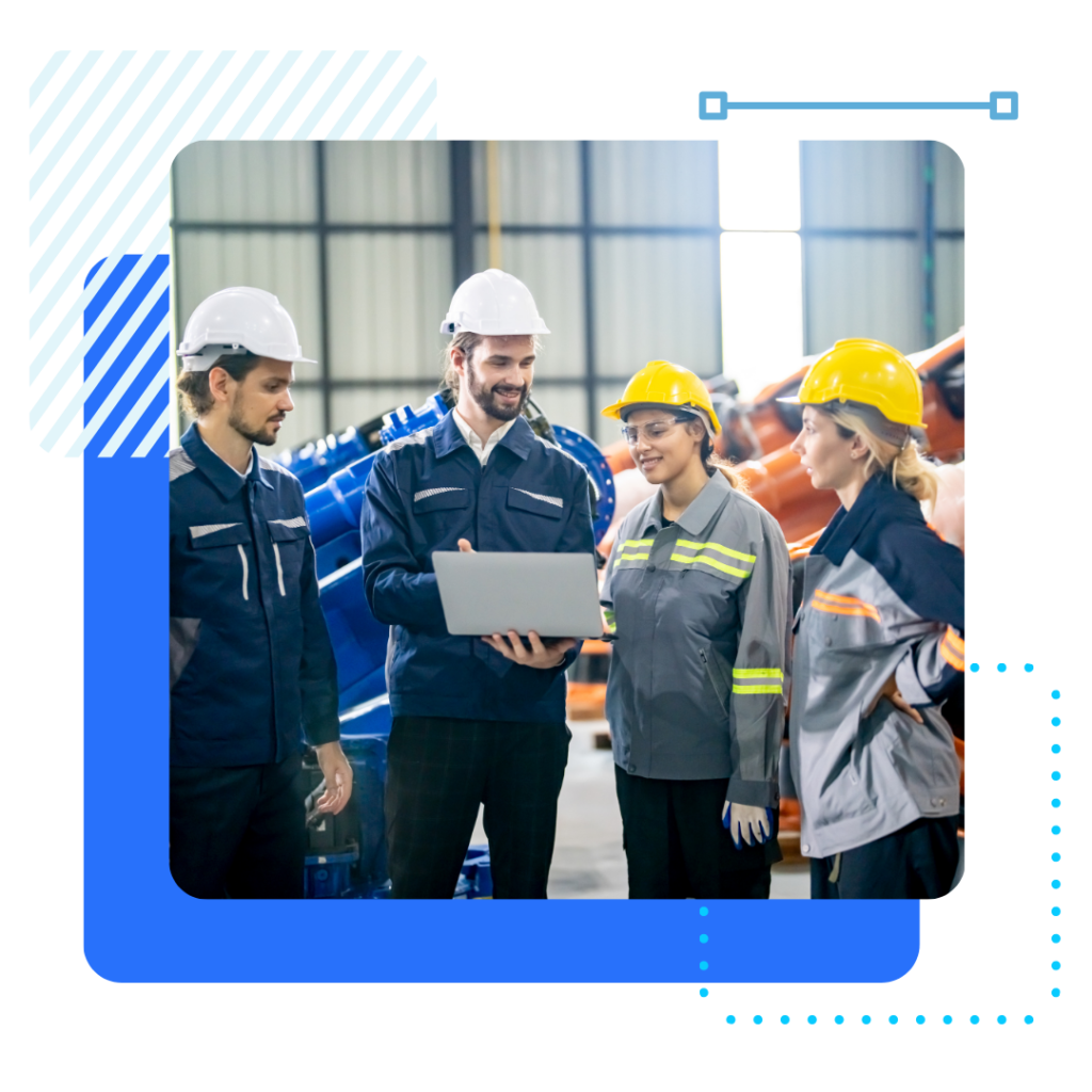 Four workers in protective gear and hard hats stand in a factory. Two look at a laptop held by a smiling colleague, discussing industrial operations.