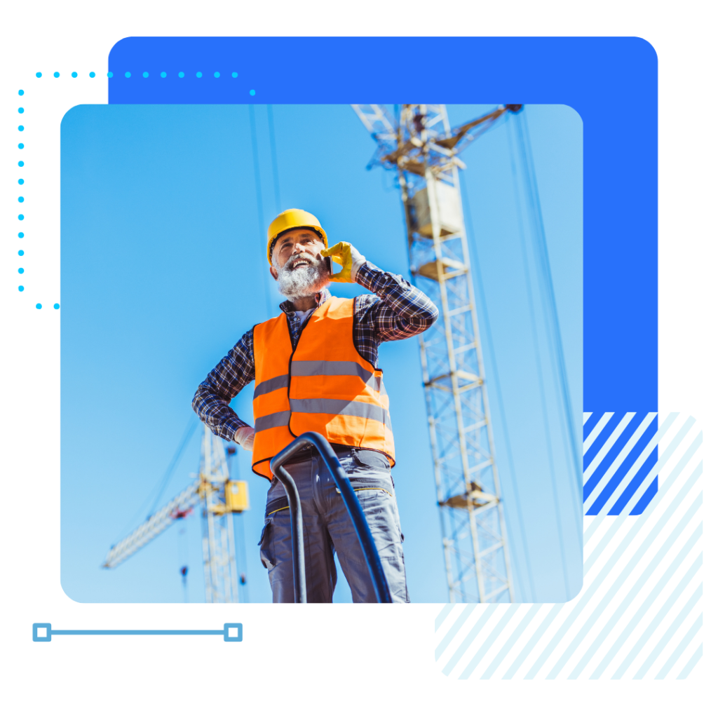 Construction worker with a beard in a hard hat and orange vest talks on a phone. Cranes in the background. Sky is clear blue, suggesting a sunny day.