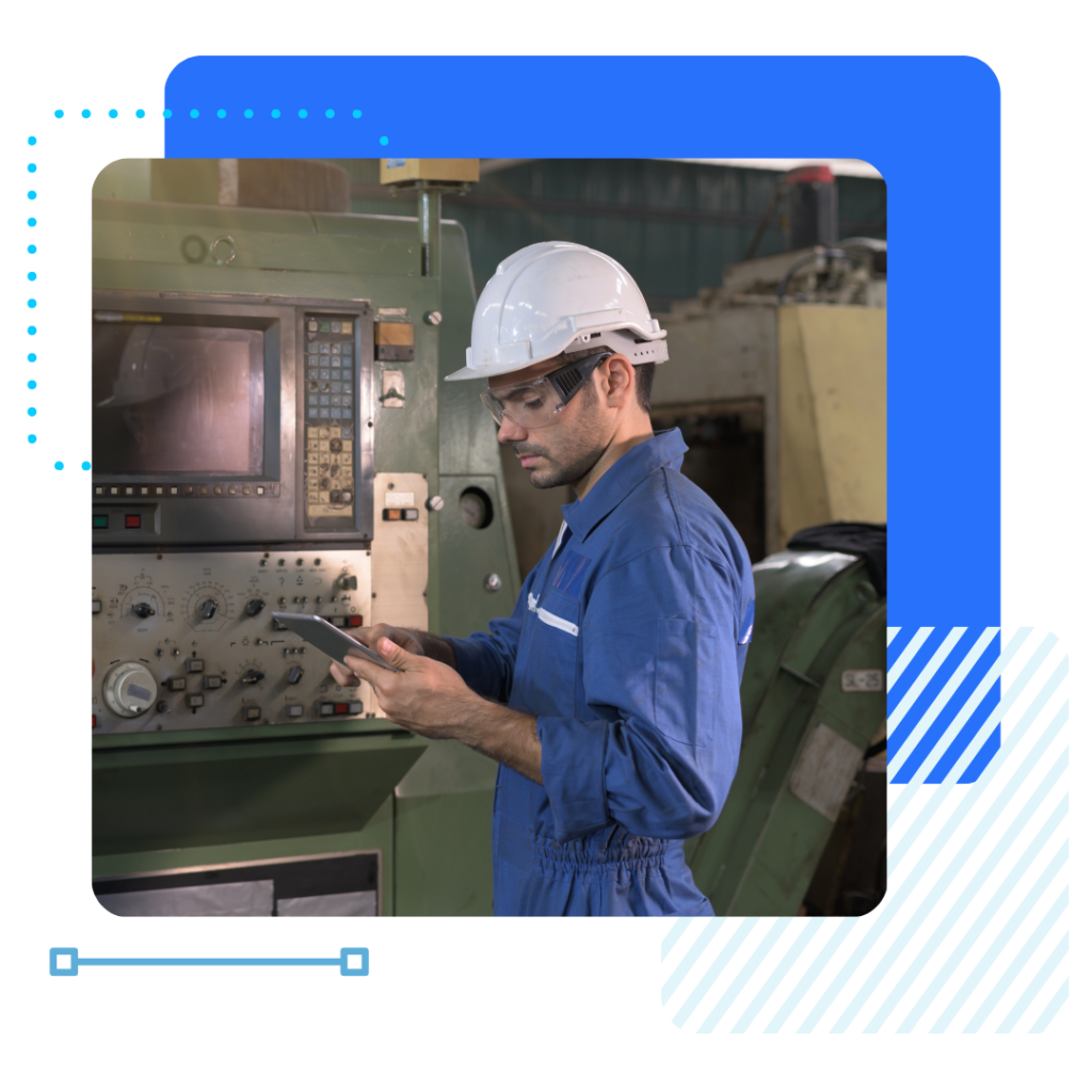 A manufacturer worker in a blue jumpsuit and white hard hat uses a tablet near industrial machinery.