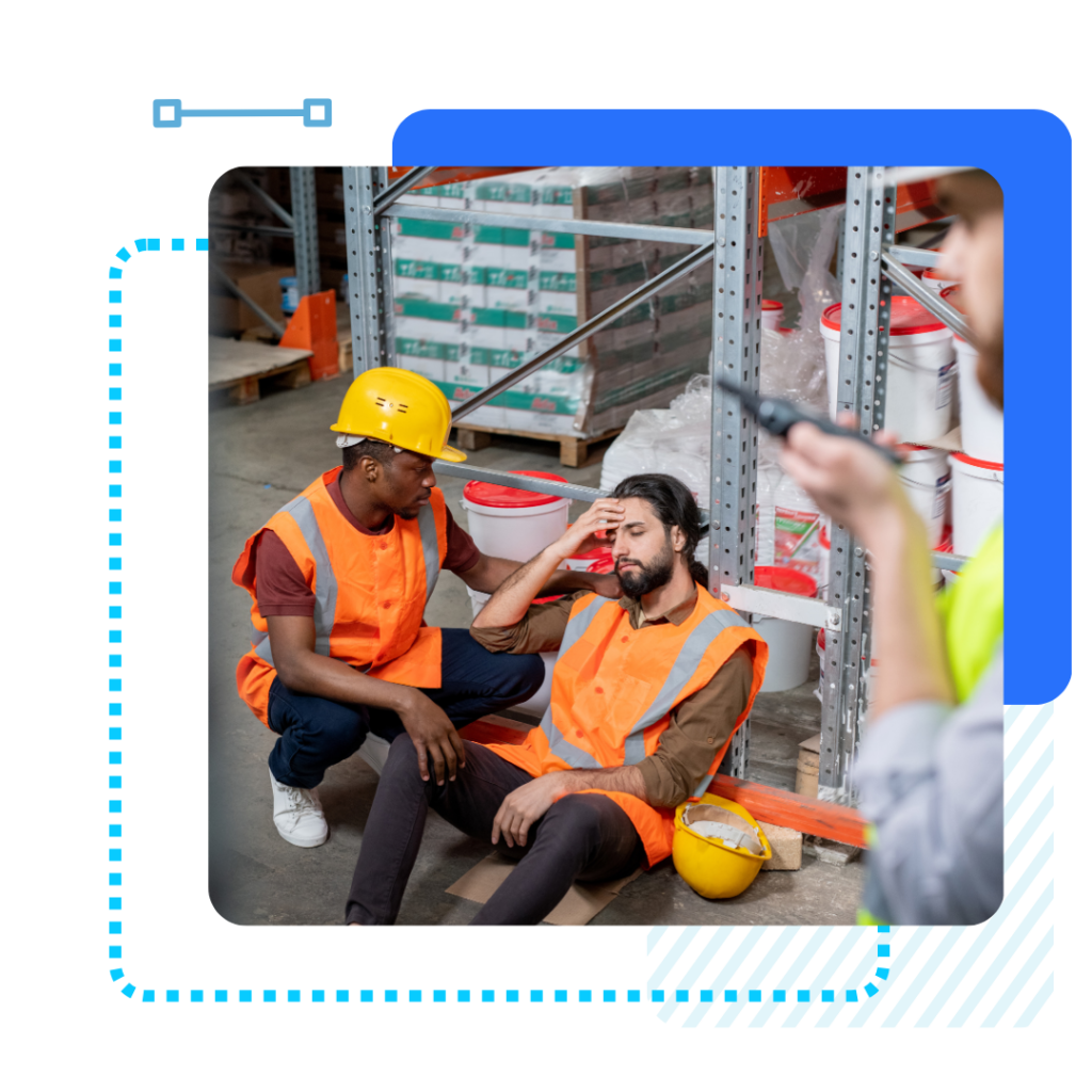 Worker sitting on the warehouse floor, looking unwell and holding his head. Another worker kneels beside him, appearing concerned. A third person uses a walkie-talkie. Orange safety vests and hard hats are worn.