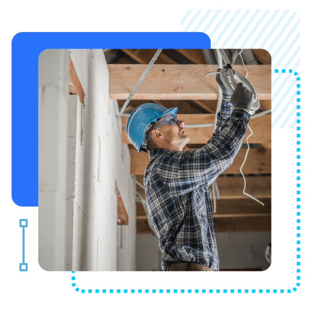 A maintenance worker in a blue hard hat and plaid shirt works on wiring in a building's ceiling.