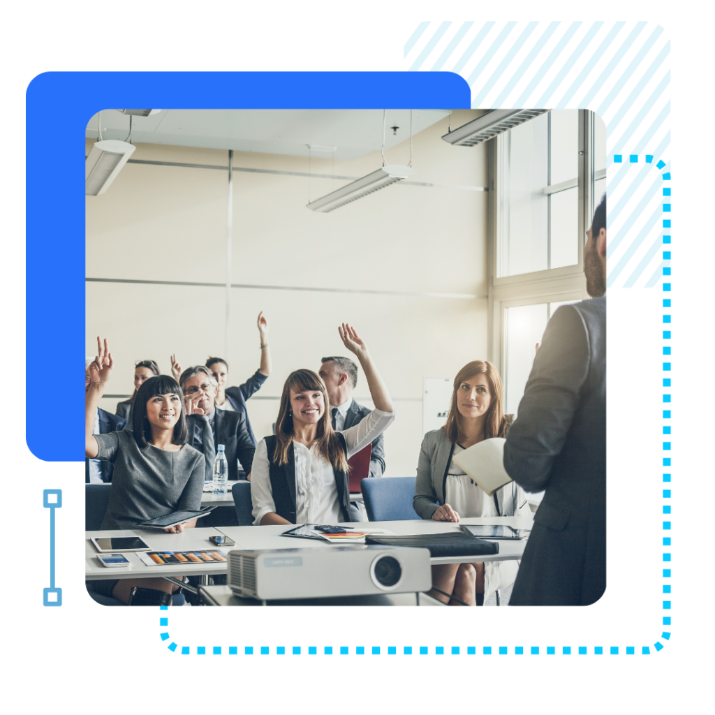 A classroom setting with adults seated at desks, some raising hands eagerly. A presenter stands in the foreground. The mood is engaged and attentive.